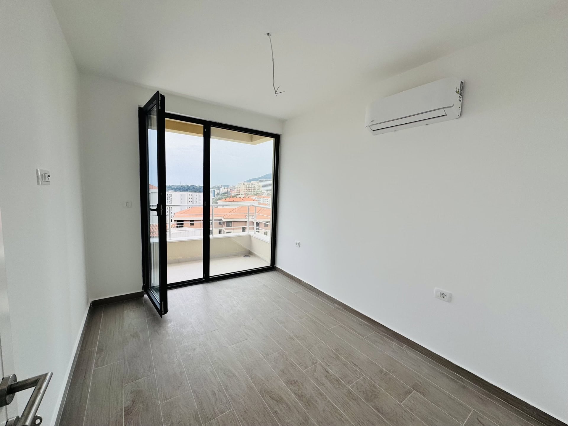 Svetla spavaća soba u stanu u Bečićima, Crna Gora: panoramski prozor, izlaz na balkon, klima-uređaj i pločice u imitaciji drveta.
