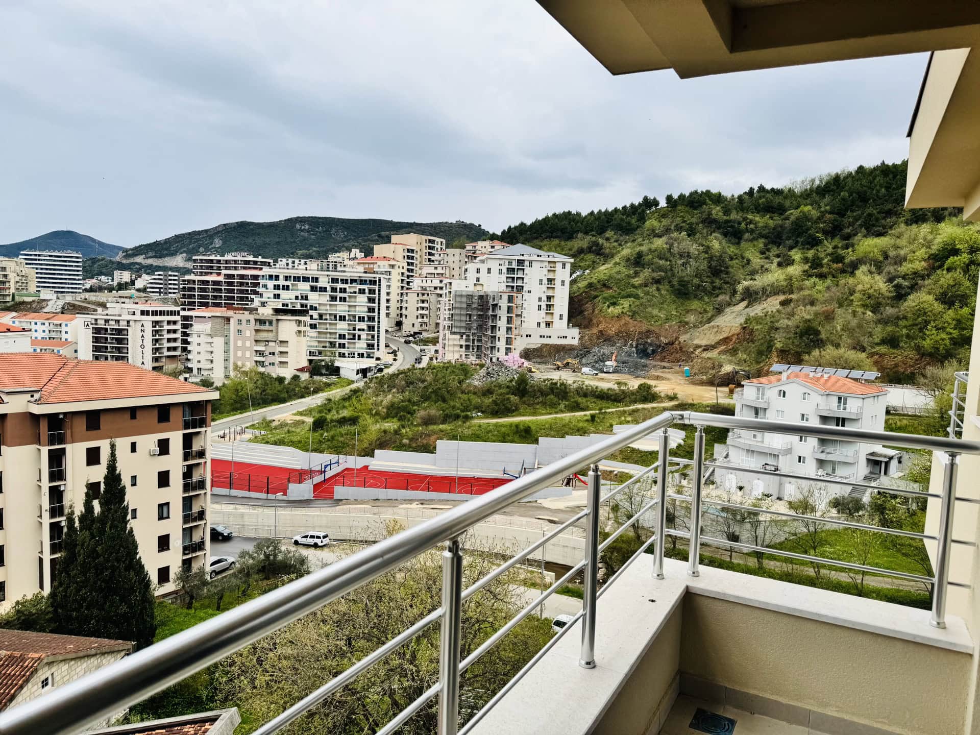 Pogled sa balkona stana u Bečićima, Crna Gora: panorama grada, stambeni kompleksi, planine i sportski teren.