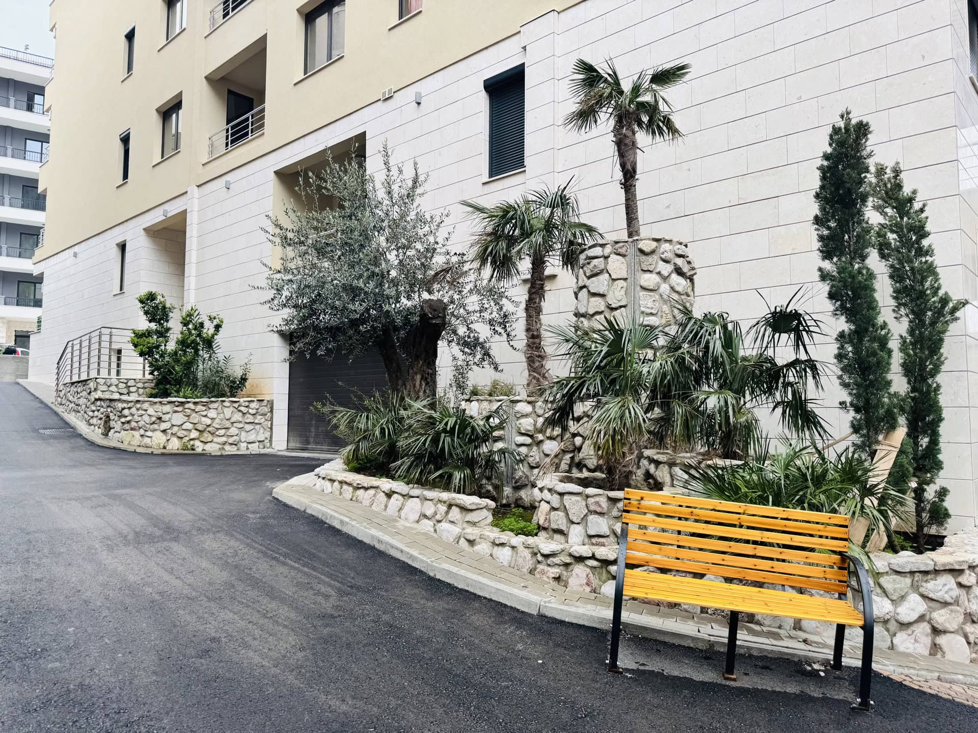 Landscaped residential complex grounds in Becici, Montenegro: stone finishes, palm trees, and a bench by the building.