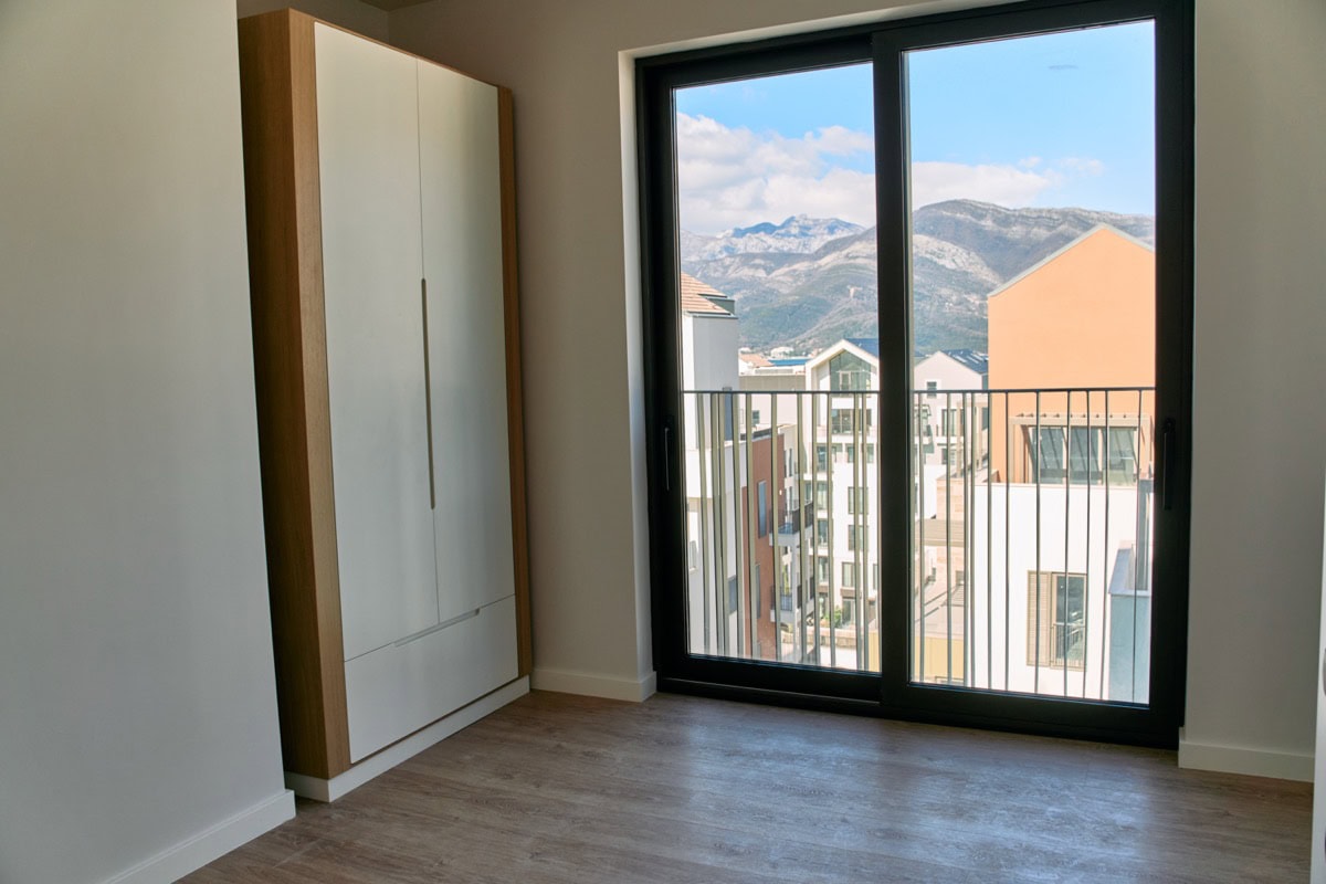 Schlafzimmer in einer Wohnung in Tivat, Montenegro: Einbauschrank, Panoramafenster mit Blick auf die Berge und moderne Wohnanlage.