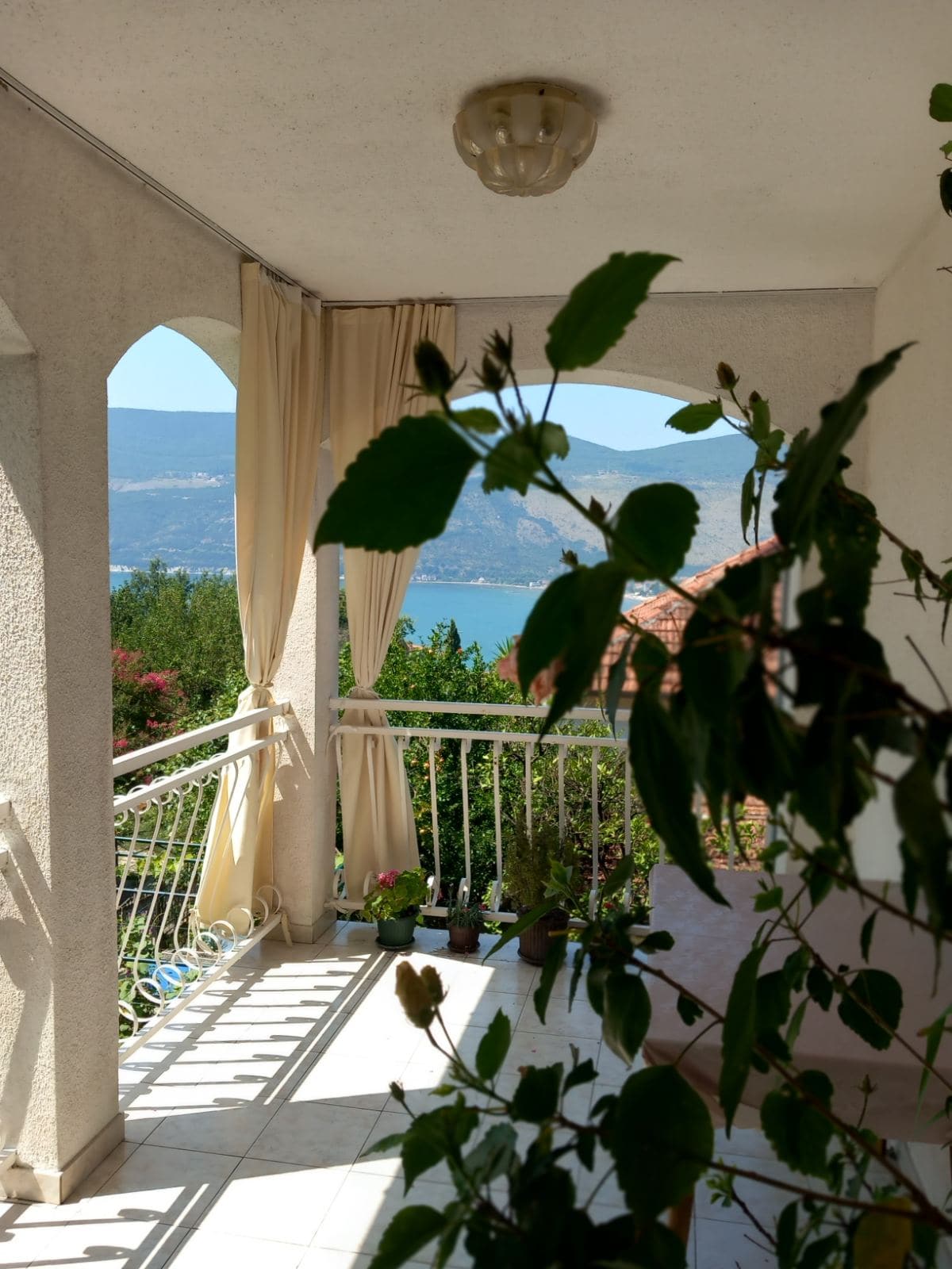 Cozy balcony with sea and mountain views in a house, Igalo, Montenegro: bright finishes, arches, and green plants.