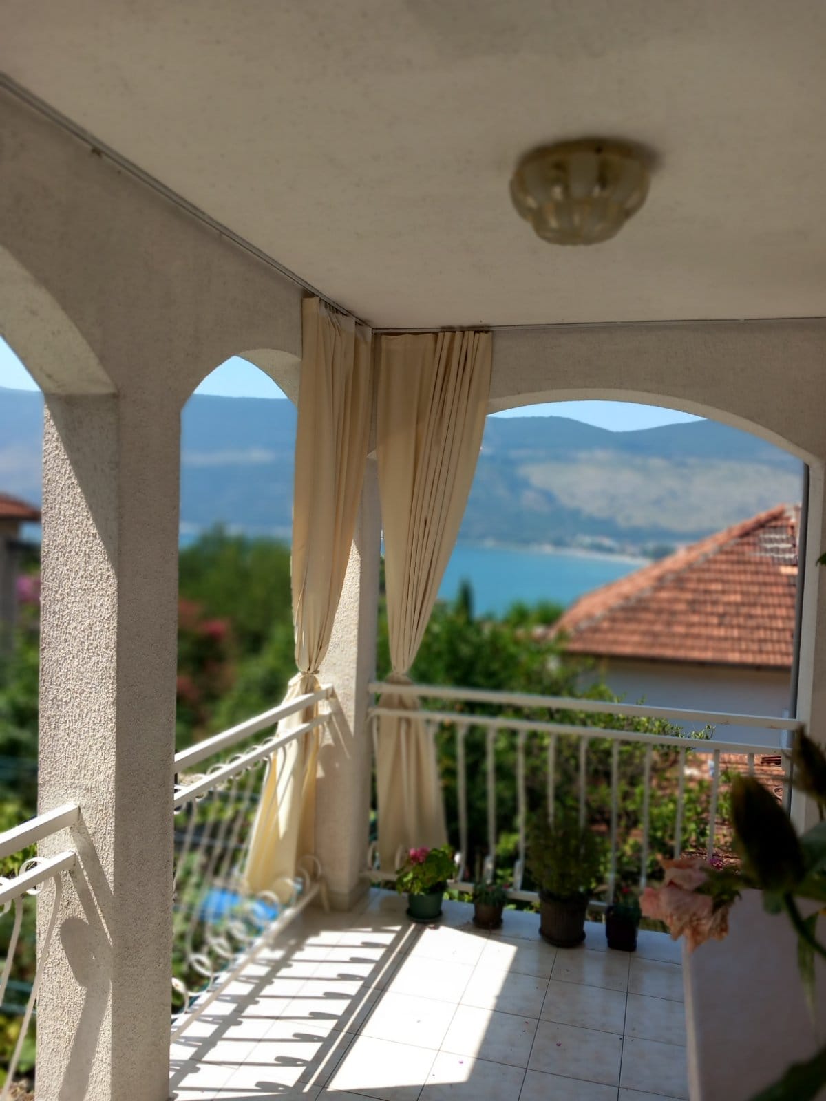 Balcony with sea and mountain views in a house in Igalo, Montenegro: arched openings, light curtains, and tiled floor.