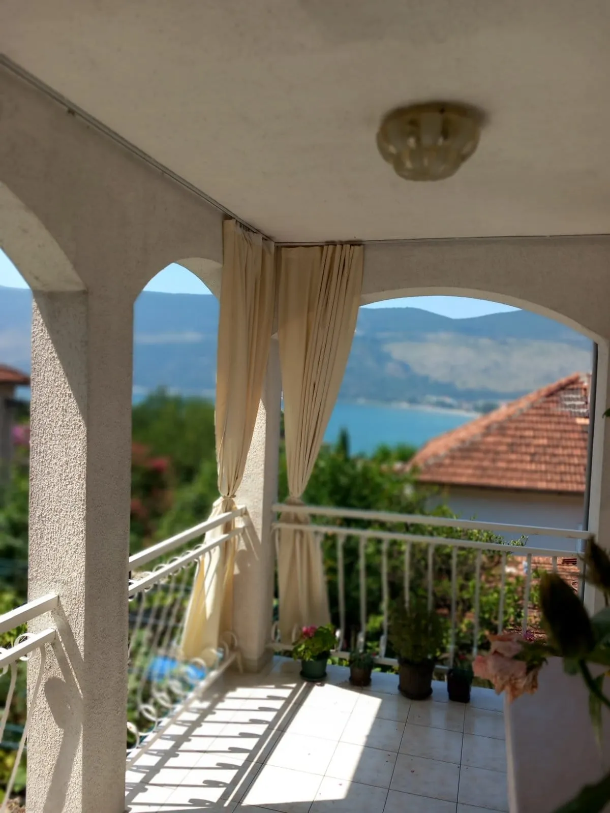 Balkon mit Blick auf das Meer und die Berge in einem Haus in Igalo, Montenegro: Rundbogenöffnungen, helle Vorhänge und Fliesenboden.
