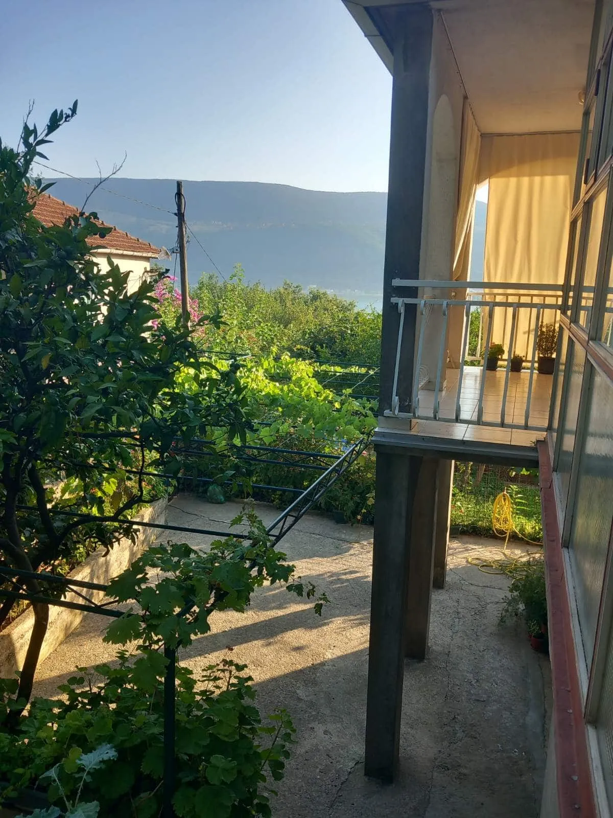 Balkon eines Hauses in Igalo, Montenegro, mit Blick auf einen grünen Garten und Berge, Terrasse mit Überdachung und Sommermöbeln.