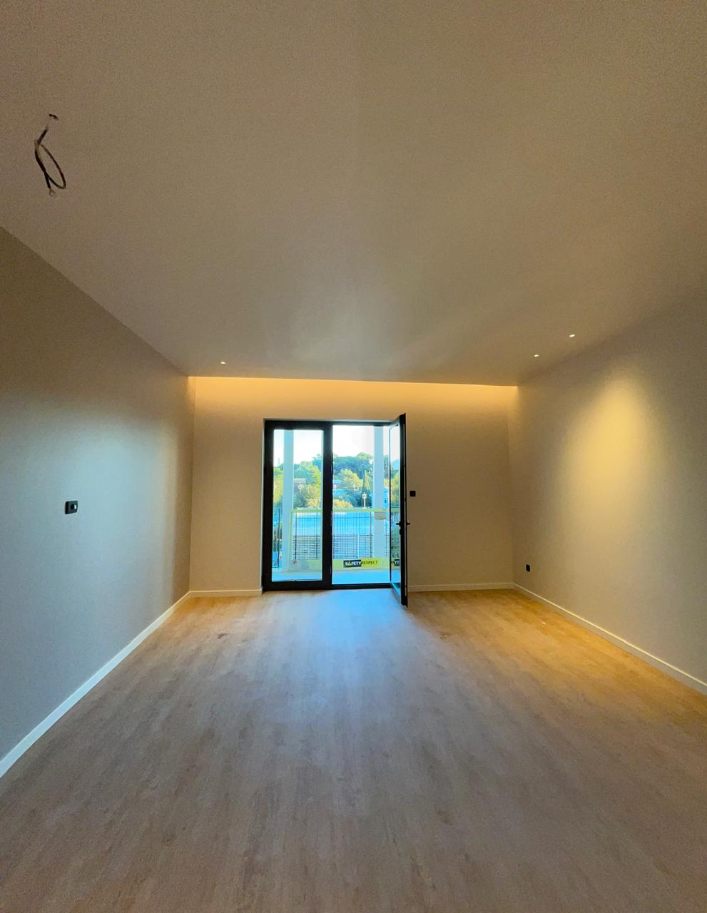 Ein hell beleuchtetes, leeres Wohnzimmer mit hellem Holzfußboden und Glastür zum Balkon.