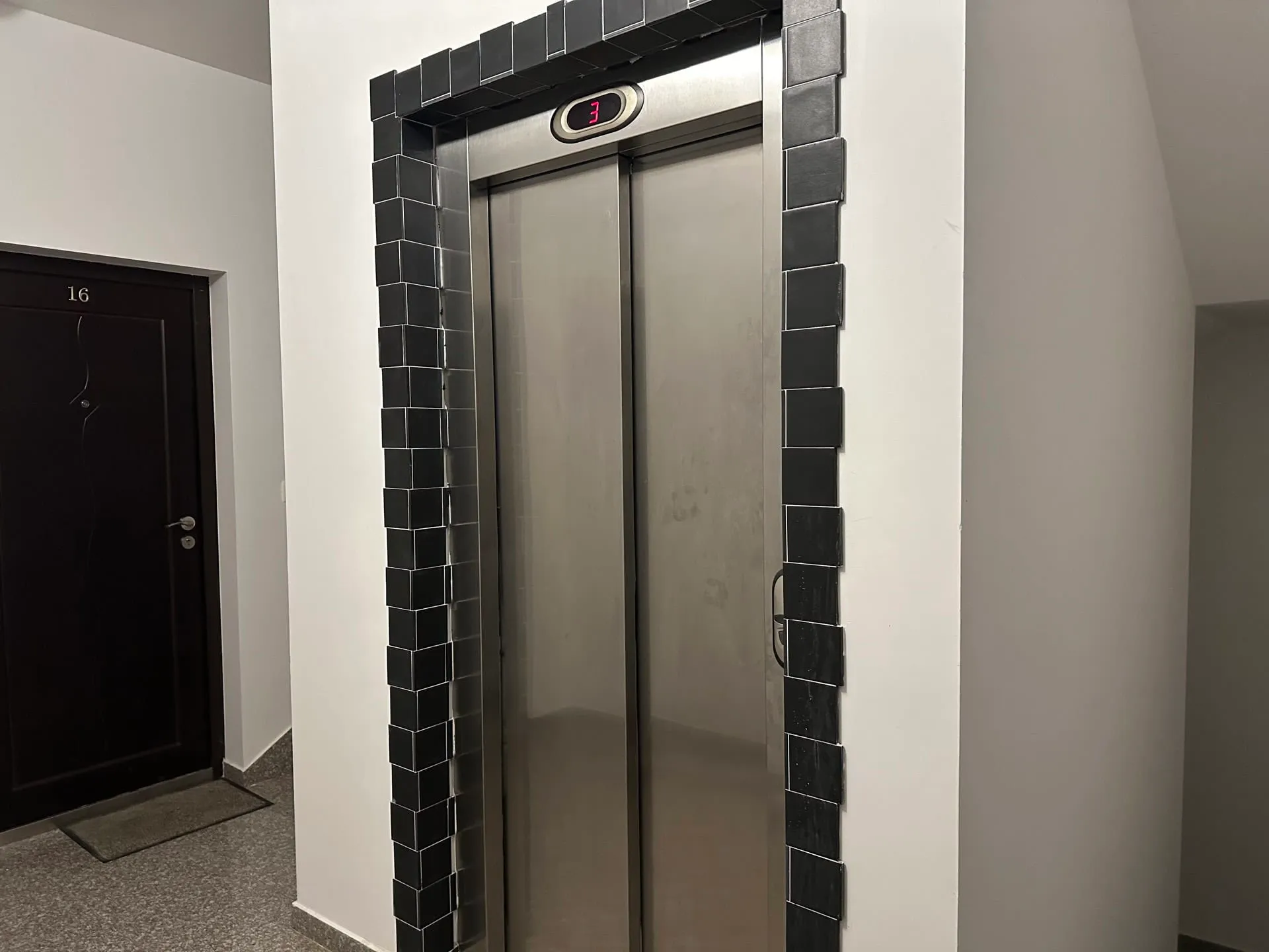 Лифт с металлическими дверями в черной tiled рамке и цифровым индикатором этажа рядом с темной дверью квартиры 16.