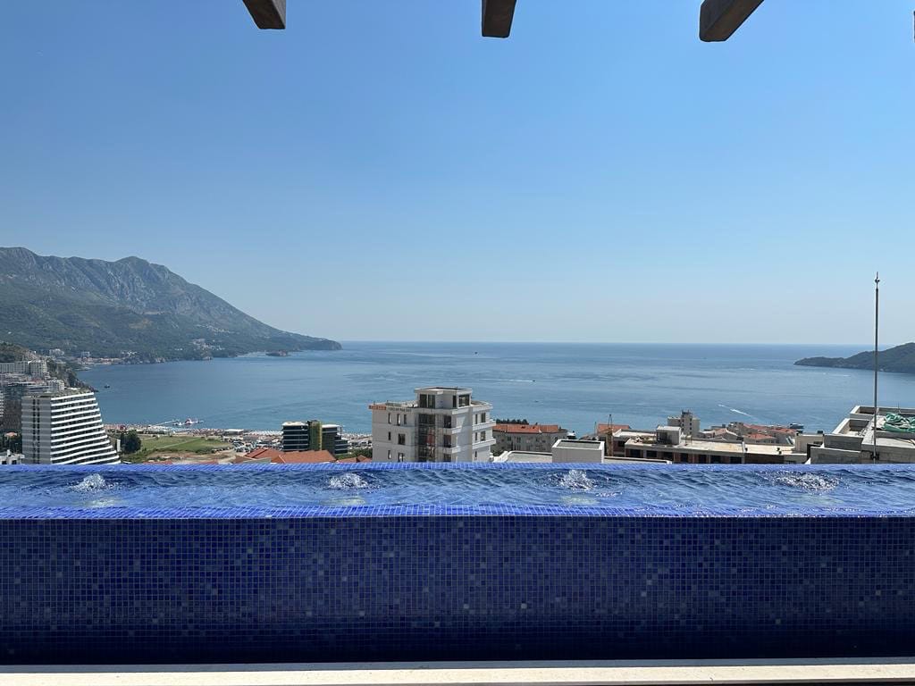 Panoramablick auf Meer und Berge vom Pool des Penthouses in Bečići, Montenegro, mit Terrasse und blauer Mosaikverkleidung.