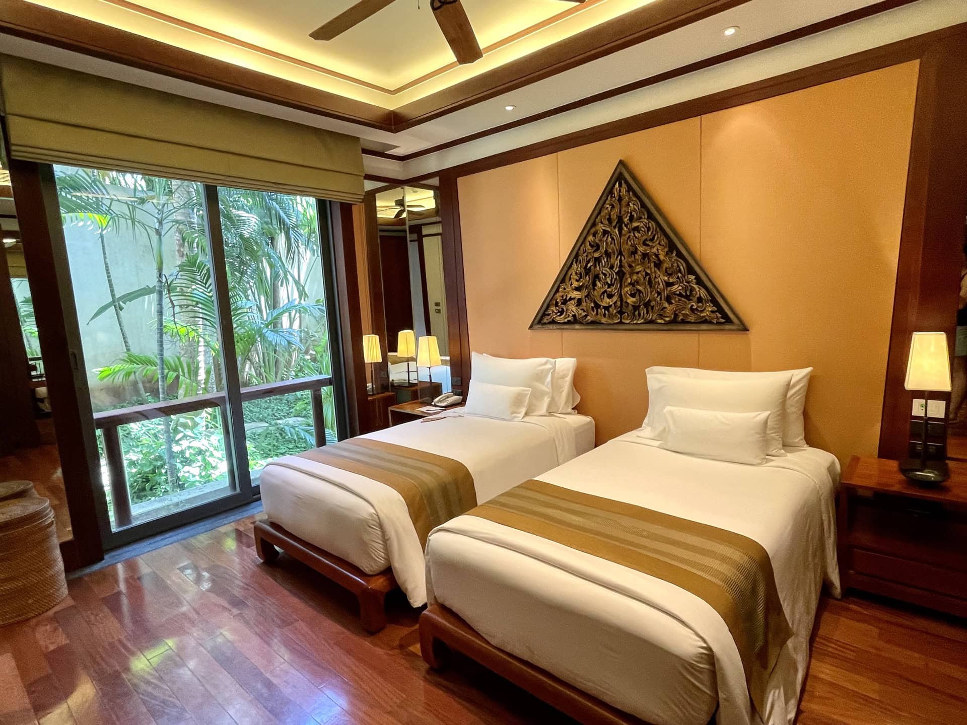 Schlafzimmer in einer Wohnung in Phuket mit zwei Betten, Holzboden, Panoramafenster und Blick auf den tropischen Garten.