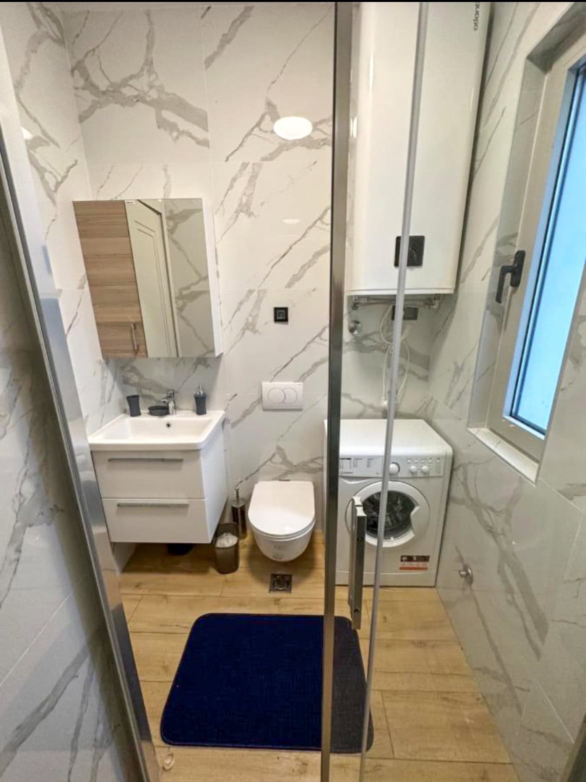 Karadağ, Budva’daki bir dairede modern banyo: mermer görünümlü fayans, duş kabini ve çamaşır makinesi.