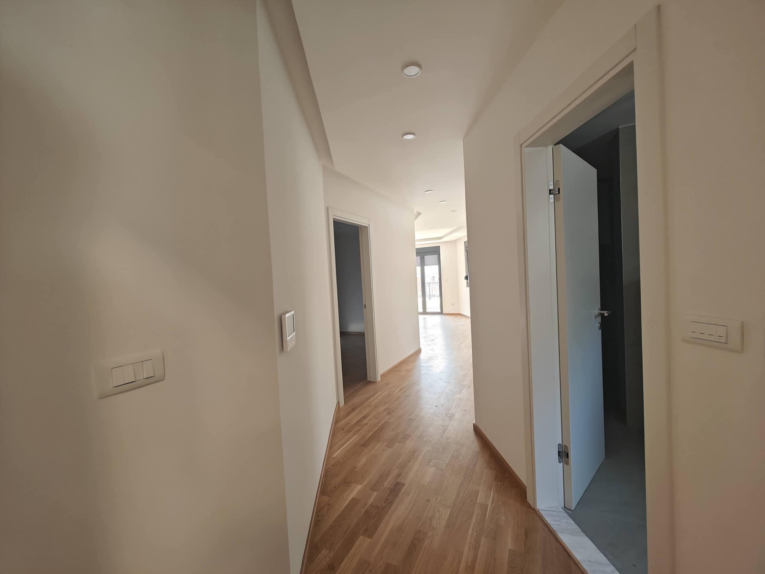 Svetao hodnik u modernom stanu u Budvi, Crna Gora: parket, beli zidovi i ugrađeno osvetljenje.