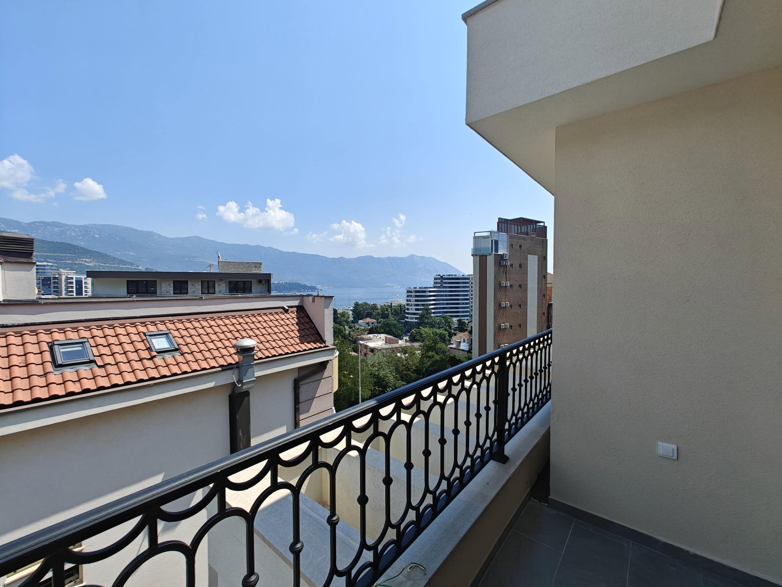 Pogled sa balkona stana u Budvi, Crna Gora: panorama grada, planina i mora, kovana ograda, terakotni krov.