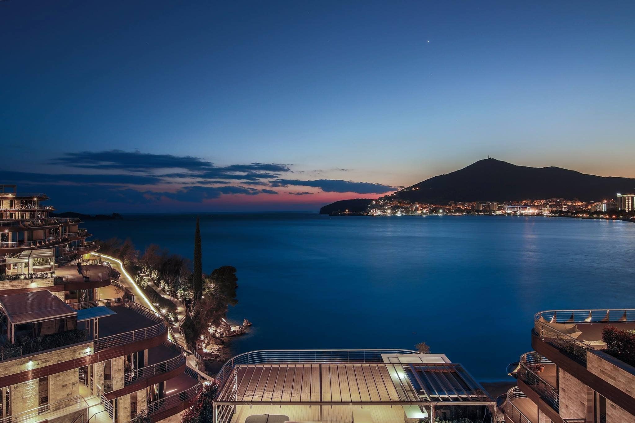 Panoramablick auf das Meer und das nächtliche Budva von einer exklusiven Wohnanlage in Montenegro mit Terrassen und Gebäudebeleuchtung.