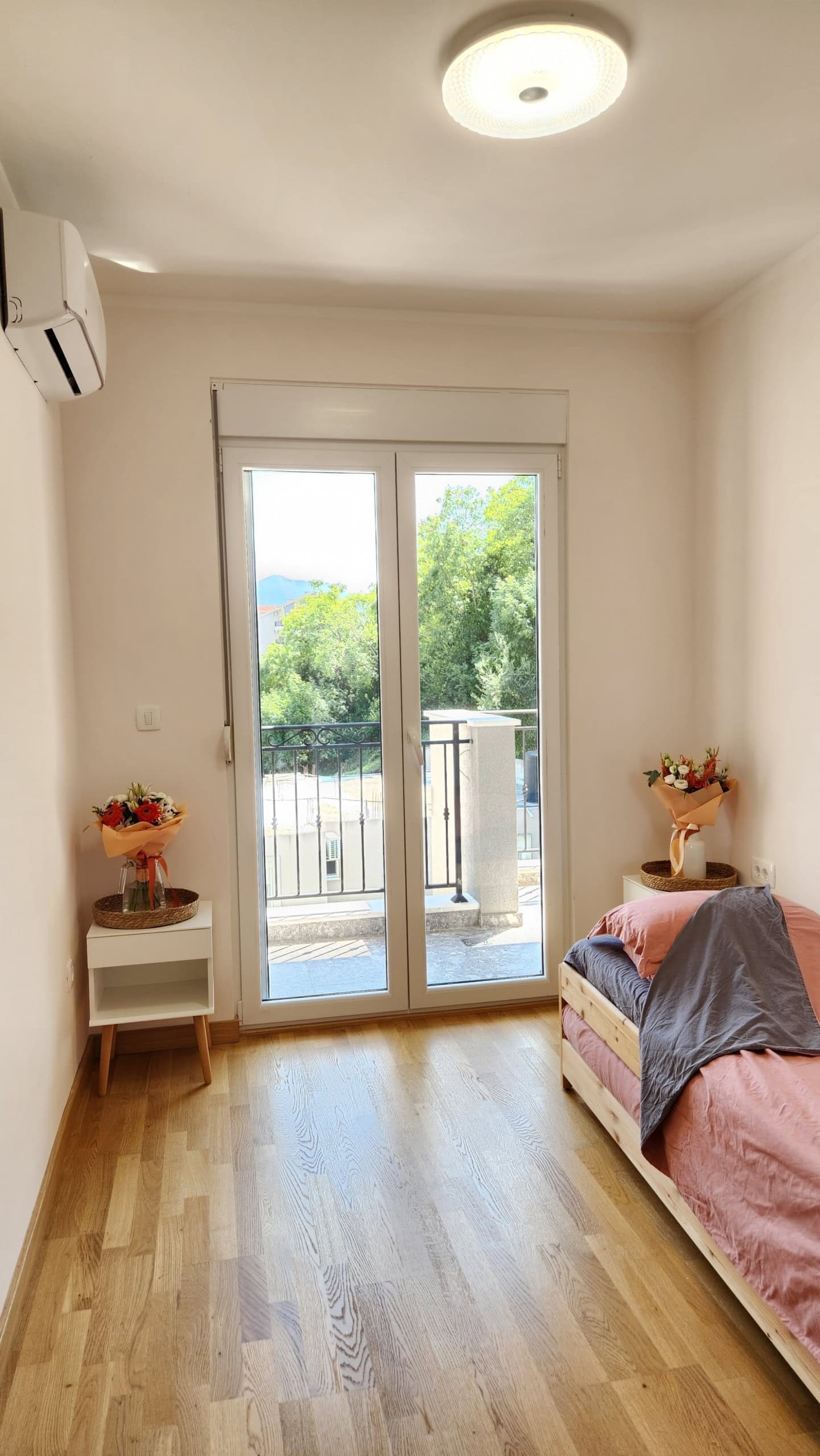 Bright bedroom in an apartment in Budva, Montenegro: bed, bedside tables, and balcony access with a view of the greenery.