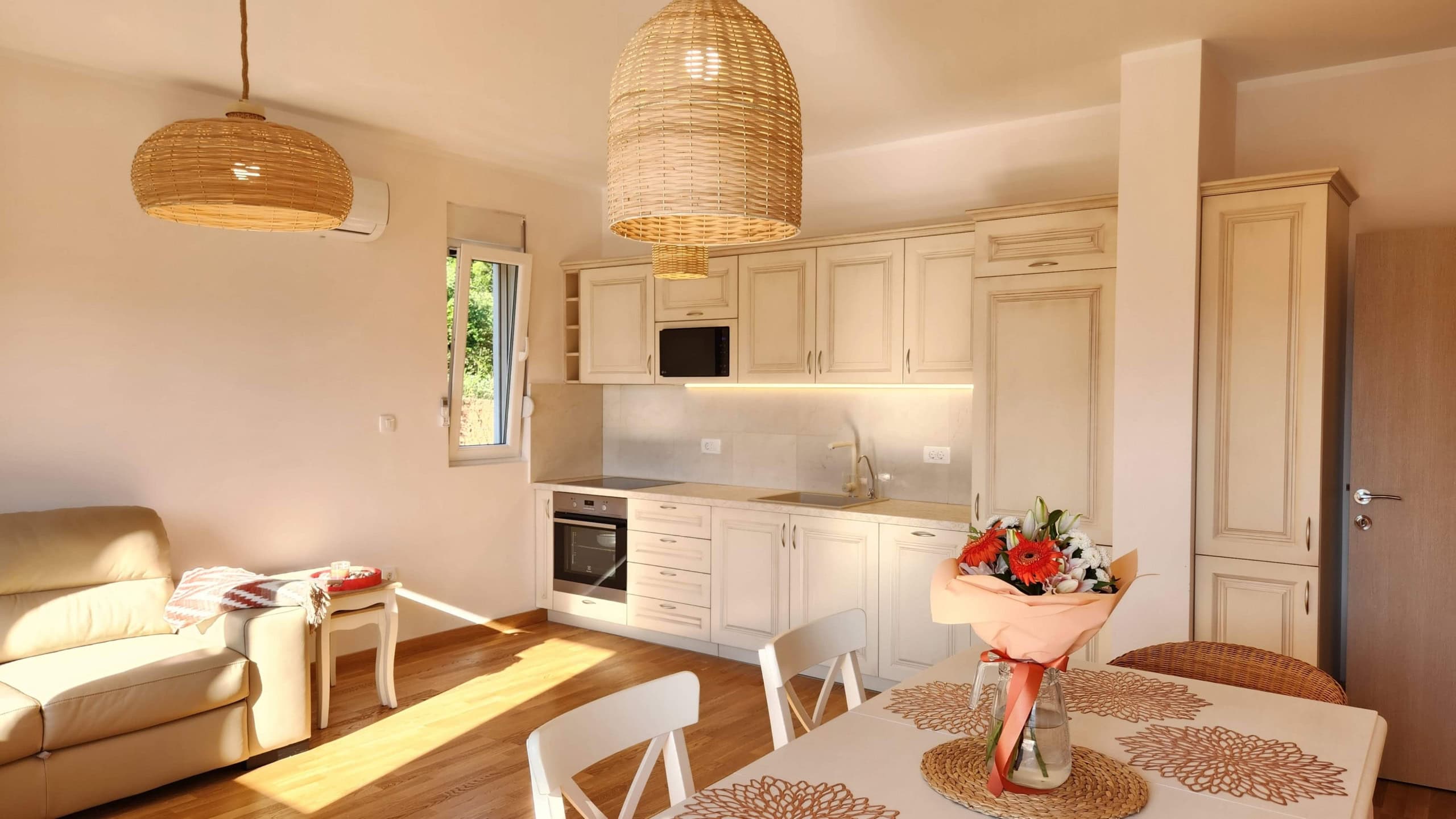 Bright kitchen-living room in a Budva apartment, Montenegro: beige cabinetry, wicker lamps, and a dining area.