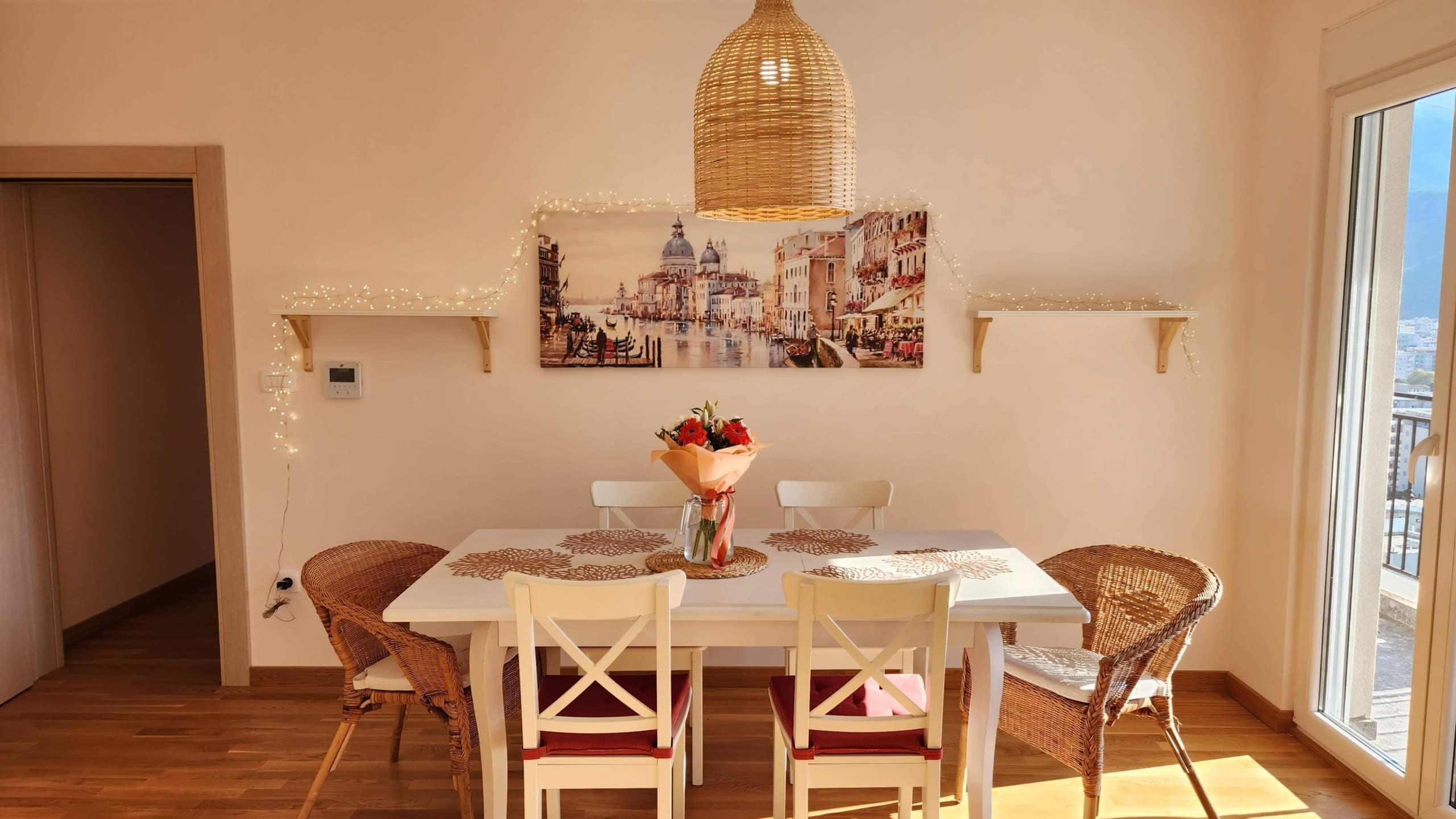 Cozy dining area in a Budva apartment, Montenegro: white table, wicker chairs, decor, and a sea view from the window.