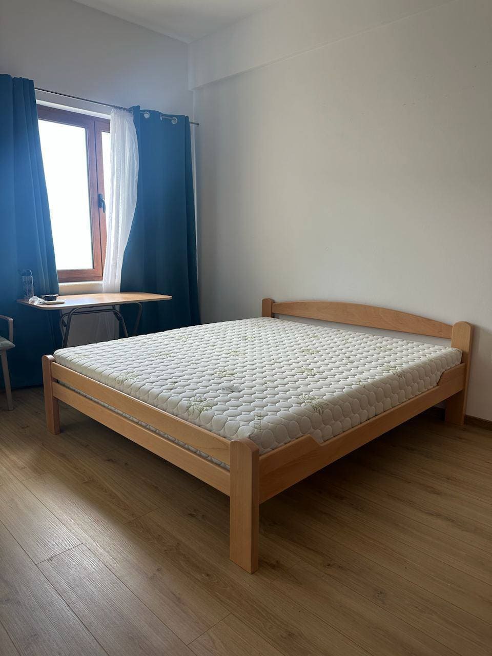 Spavaća soba sa bračnim krevetom i svetlim laminatom u stanu u Bečićima, Crna Gora. Prozor sa plavim zavesama.