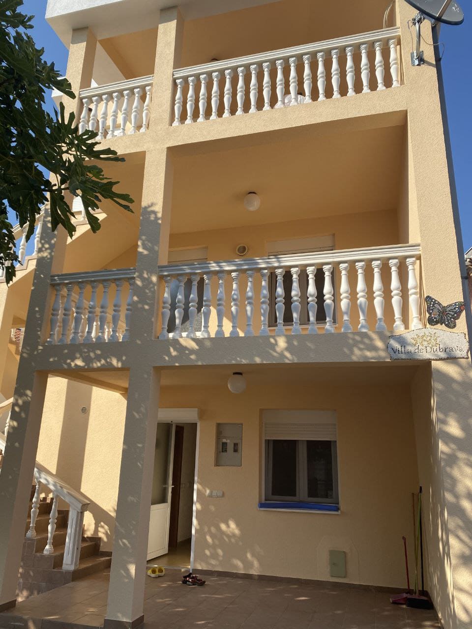 Фасад трехэтажной виллы Villa de Dubrava в Добра Вода, Черногория, с открытыми террасами и белыми балюстрадами.