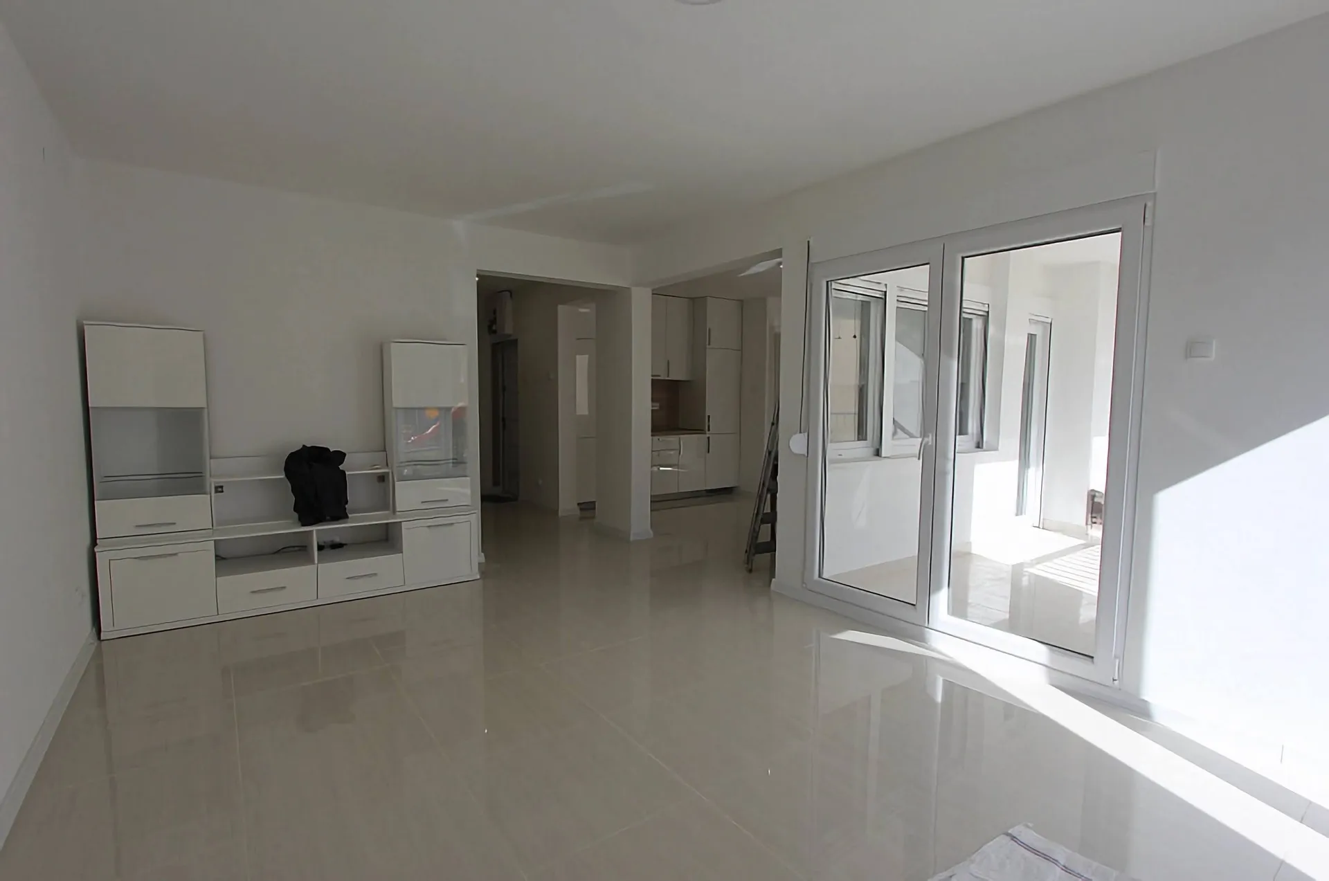 Ein modernes unmöbliertes Wohnzimmer mit weißen Wänden, einer weißen modularen Schrankwand und großen Glastüren zum Balkon.