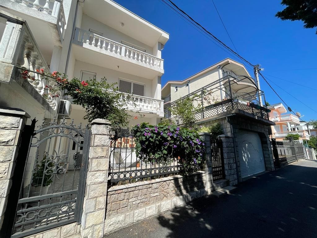 Dreistöckiges Haus in Bar, Montenegro: Fassade mit weißen Balkonen, Steinzaun und blühende Pflanzen am Eingang.