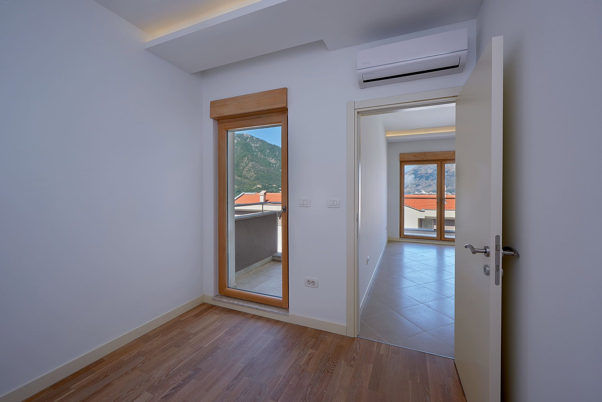 Bright bedroom in an apartment in Dobrota, Montenegro: laminate flooring, access to a balcony with mountain views, and a modern air conditioner.
