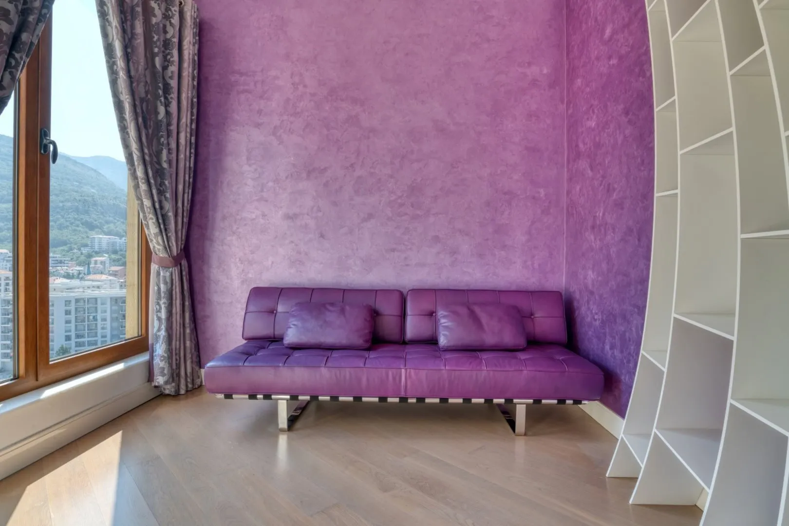 Fioletowa sofa przy oknie z widokiem na góry, fioletowa ściana i biała rega.