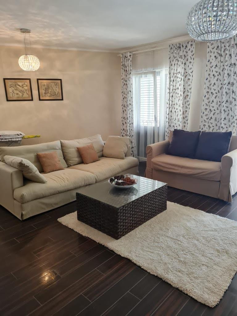 Przytulny salon w mieszkaniu w Petrovacu, Czarnogóra: jasne sofy, pleciony stolik kawowy i ciemna podłoga.