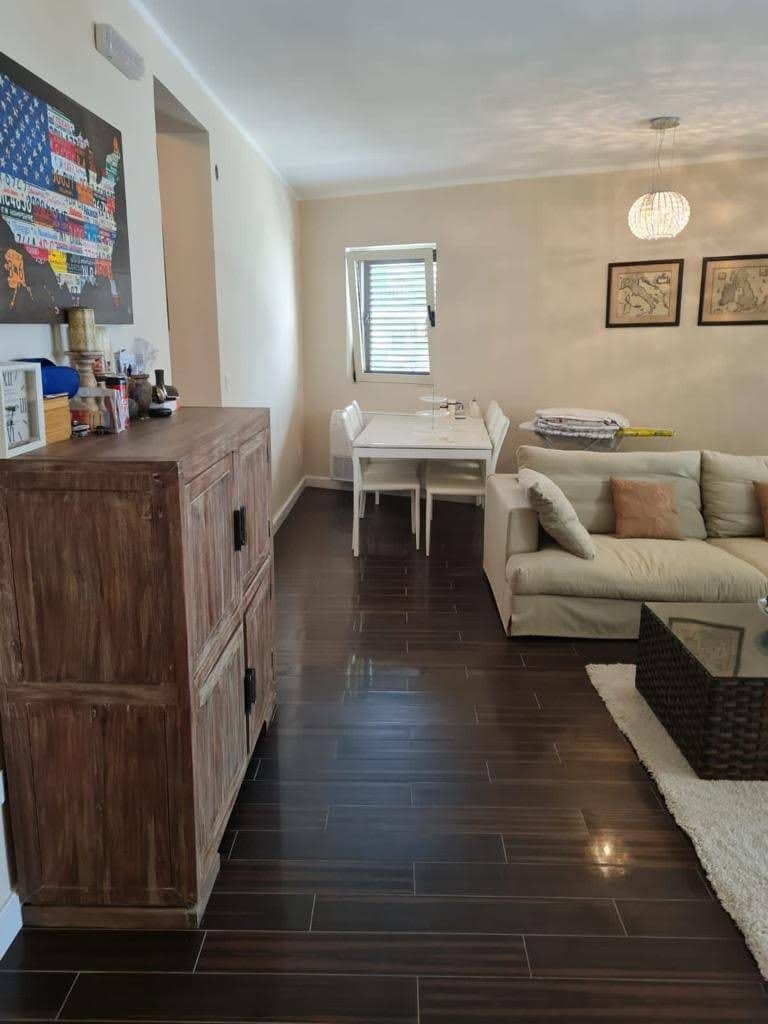 Salon w mieszkaniu w Petrovacu, Czarnogóra: jasna sofa, jadalnia, drewniana komoda i ciemna podłoga z płytek.