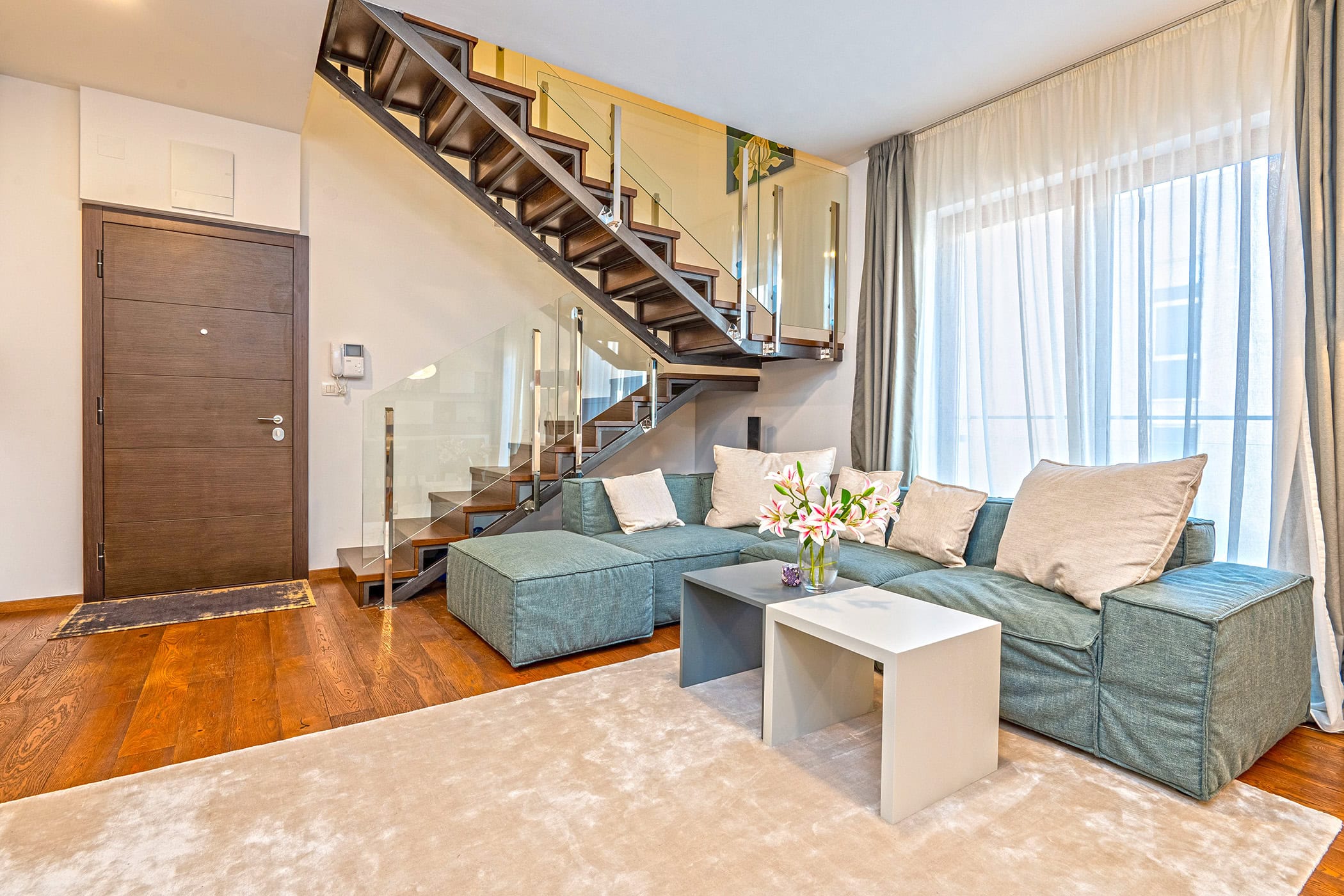 Wohnzimmer in einem Duplex in Budva, Montenegro: moderne Treppe mit Glasgeländer, Sofa und Parkettboden.