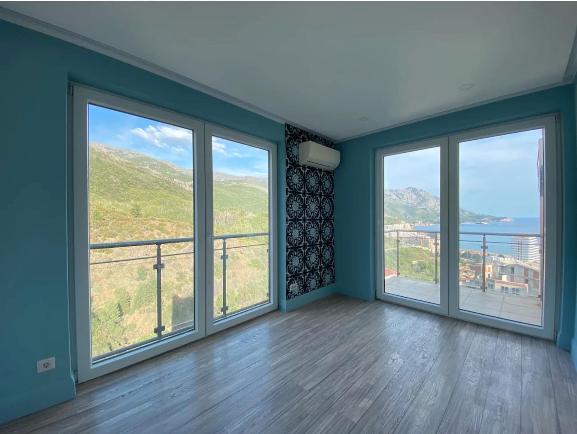 Wohnzimmer in einer Wohnung in Bečići, Montenegro: Panoramafenster mit Blick auf das Meer und die Berge, Laminatboden und Akzentwand.