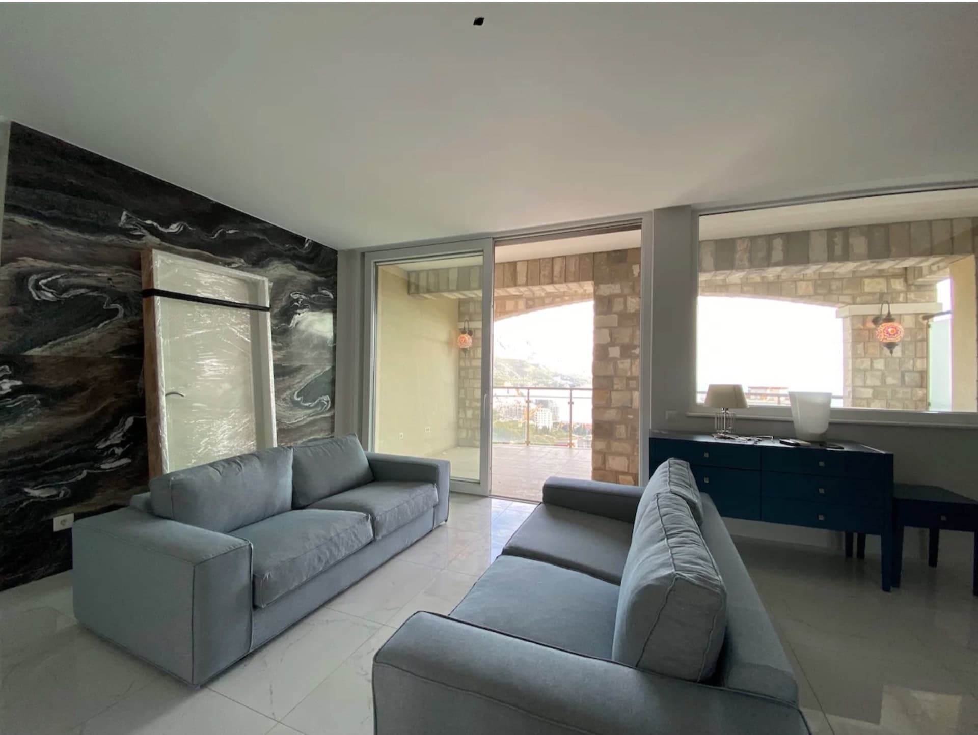 Wohnzimmer in einer Wohnung in Bečići, Montenegro: graue Sofas, eine Akzentwand aus Stein und Zugang zur Terrasse mit Meerblick.