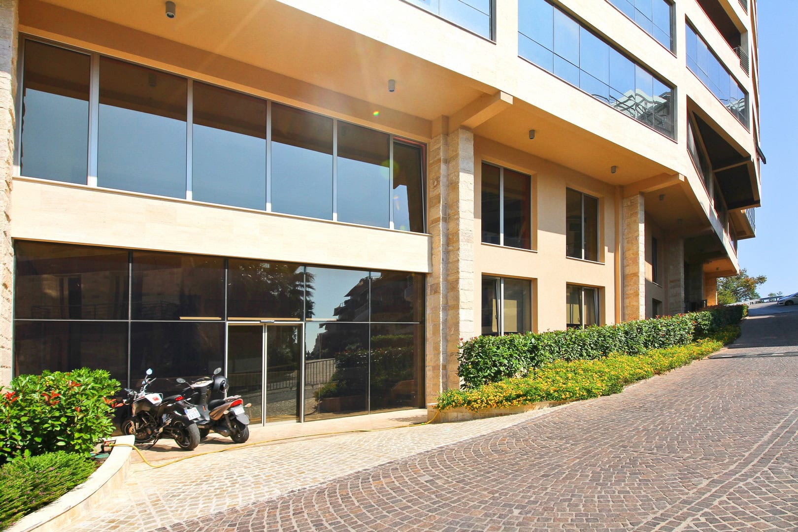 Fassade des modernen Businesscenters Harmonia in Budva, Montenegro, mit Panoramaverglasung und gepflasterter Zufahrtsstraße.