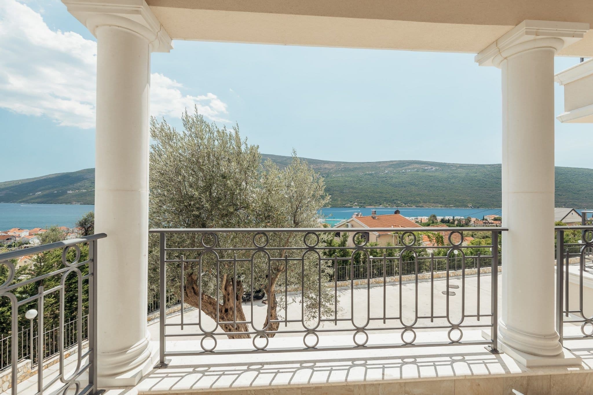Blick vom Balkon mit Säulen auf das Meer und die Berge in einer Wohnanlage, Kumbor, Montenegro. Schmiedeeisernes Geländer und Olivenbaum.