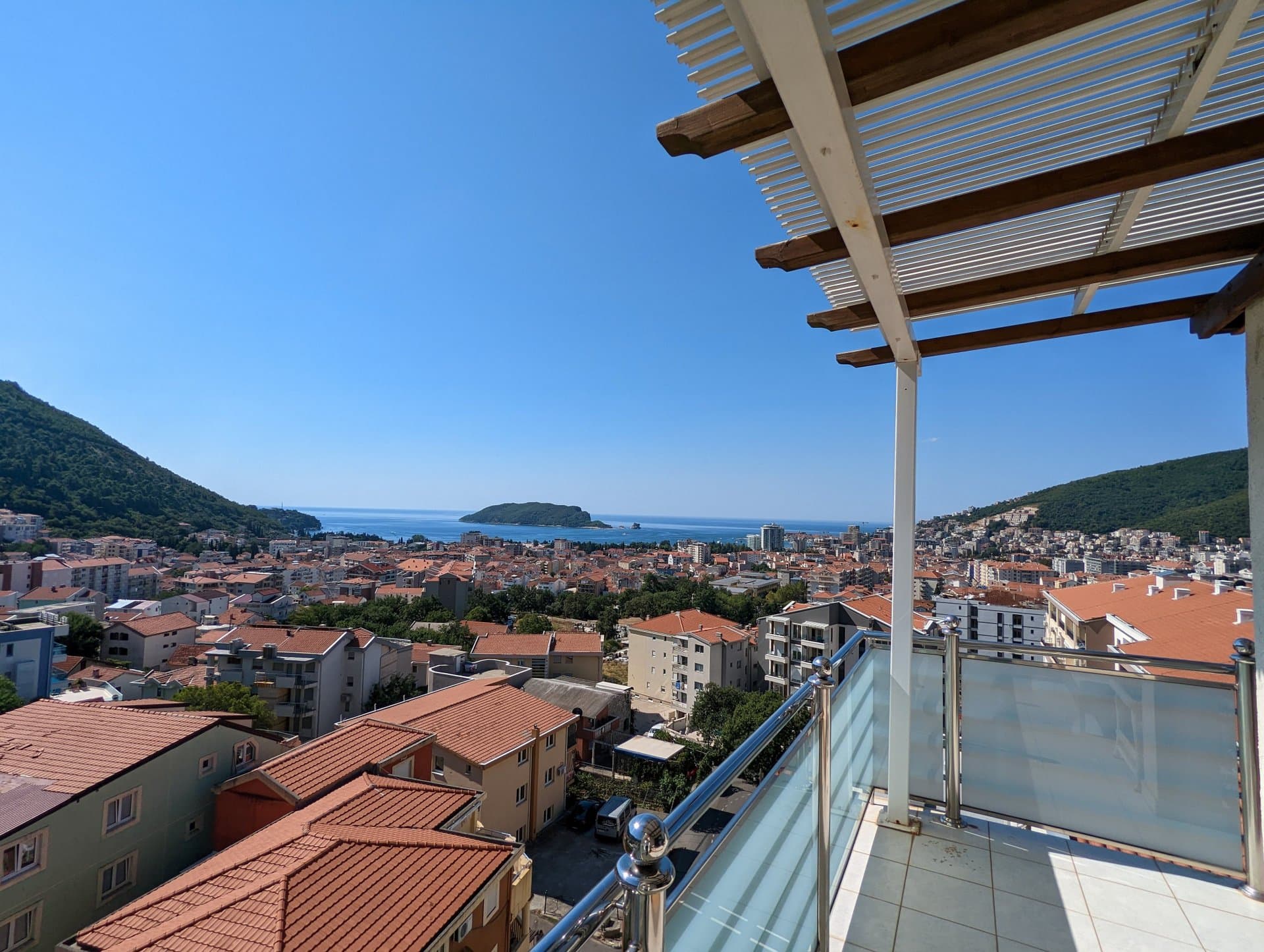 Panoramablick auf das Meer und Budva von der Terrasse des Penthouses in Montenegro, Glasgeländer und Überdachung, sonniger Tag.