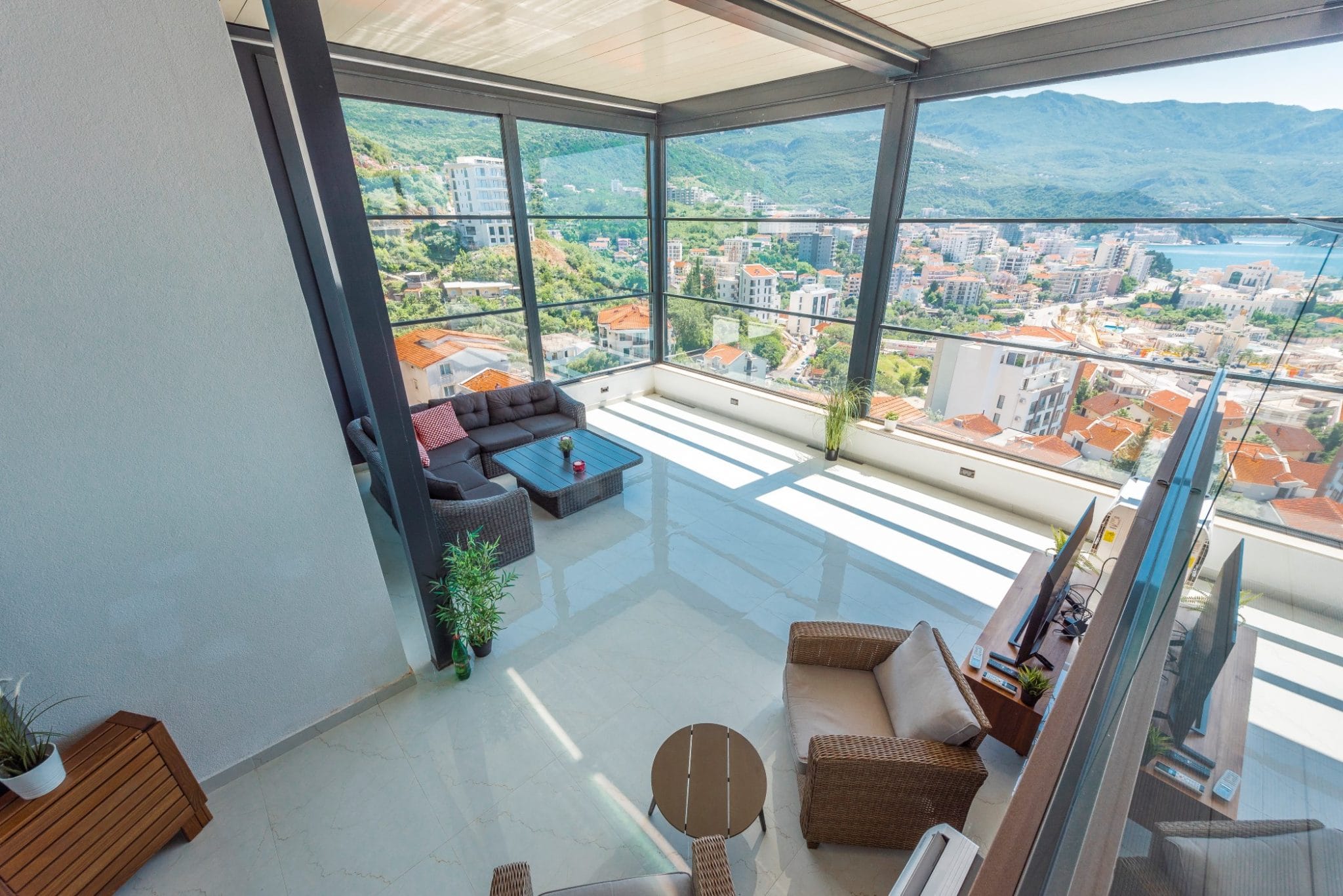 Wohnzimmer im Penthouse in Bečići, Montenegro: Panoramaverglasung, geflochtene Möbel und Blick auf das Meer und die Berge.