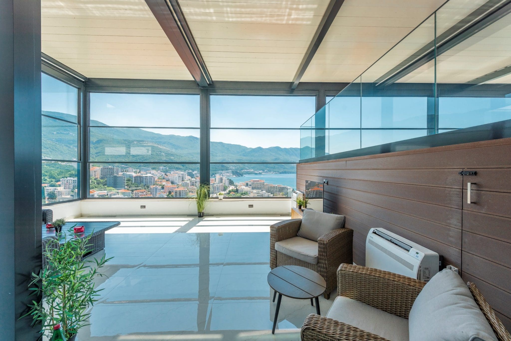 Penthouse-Terrasse in Bečići, Montenegro: Panoramaverglasung, geflochtene Möbel und Blick auf das Meer und die Berge.