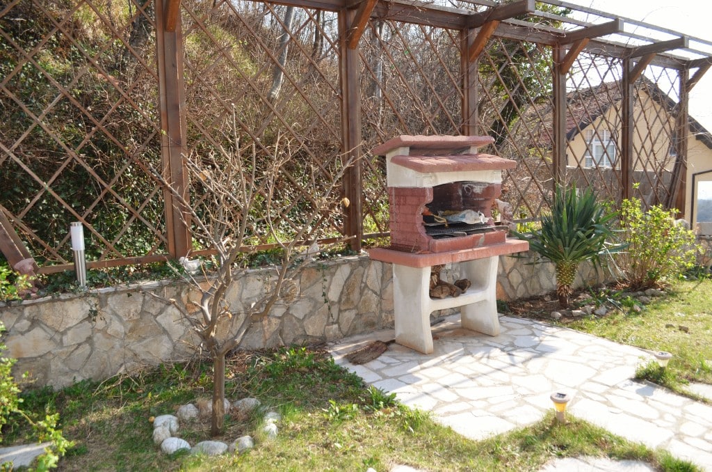 Karadağ’ın Bar şehrindeki daire terasında taş ızgaralı barbekü alanı, ahşap pergola ve bahçe.