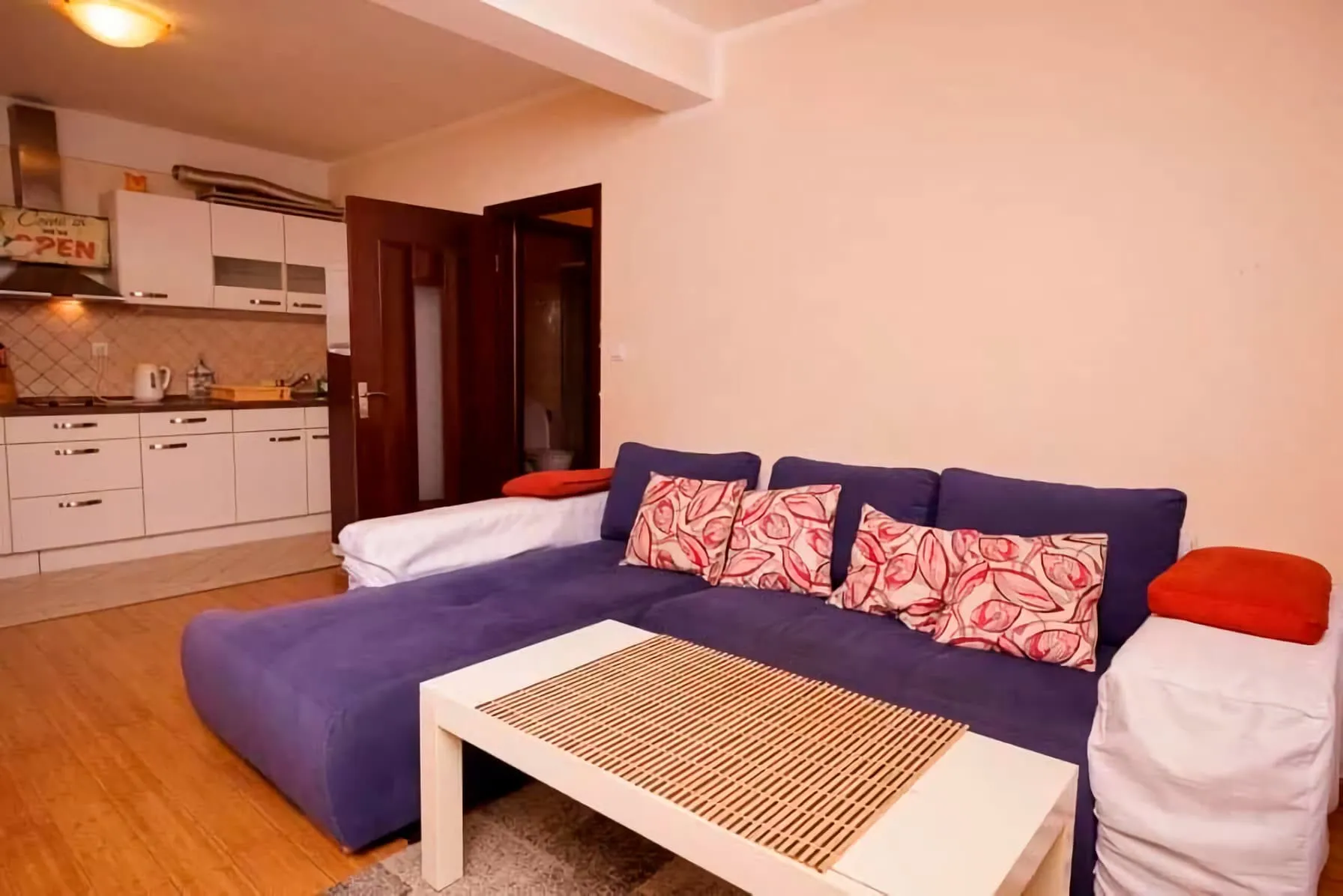 Dnevna soba sa plavom ugaonom garniturom i kuhinjom u stanu, Budva, Crna Gora. Moderan enterijer sa belim nameštajem.