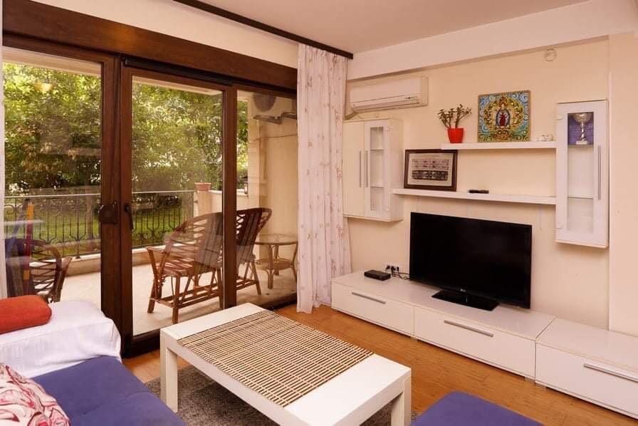Wohnzimmer mit Zugang zur Terrasse in einer Wohnung in Budva, Montenegro: helle Möbel, TV-Bereich und Blick auf die Grünanlage.