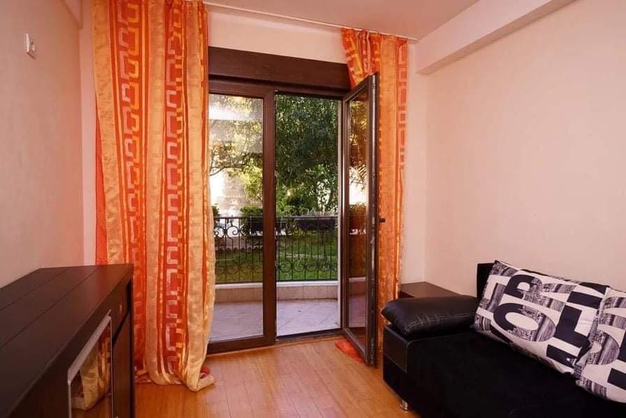 Wohnzimmer in einer Wohnung in Budva, Montenegro: schwarzes Sofa, Parkettboden, Zugang zum Balkon mit Blick auf den grünen Garten.