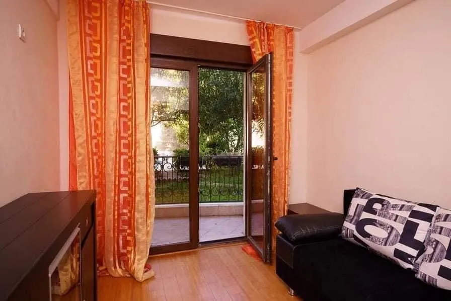 Dnevna soba u stanu u Budvi, Crna Gora: crni kauč, parketni pod, izlaz na balkon s pogledom na zelenu baštu.
