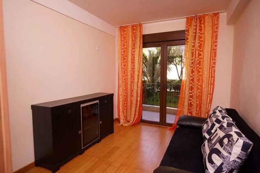 Wohnzimmer in einer Wohnung in Budva, Montenegro: schwarzes Sofa, Kommode, helle Wände und Zugang zum Balkon mit Blick ins Grüne.