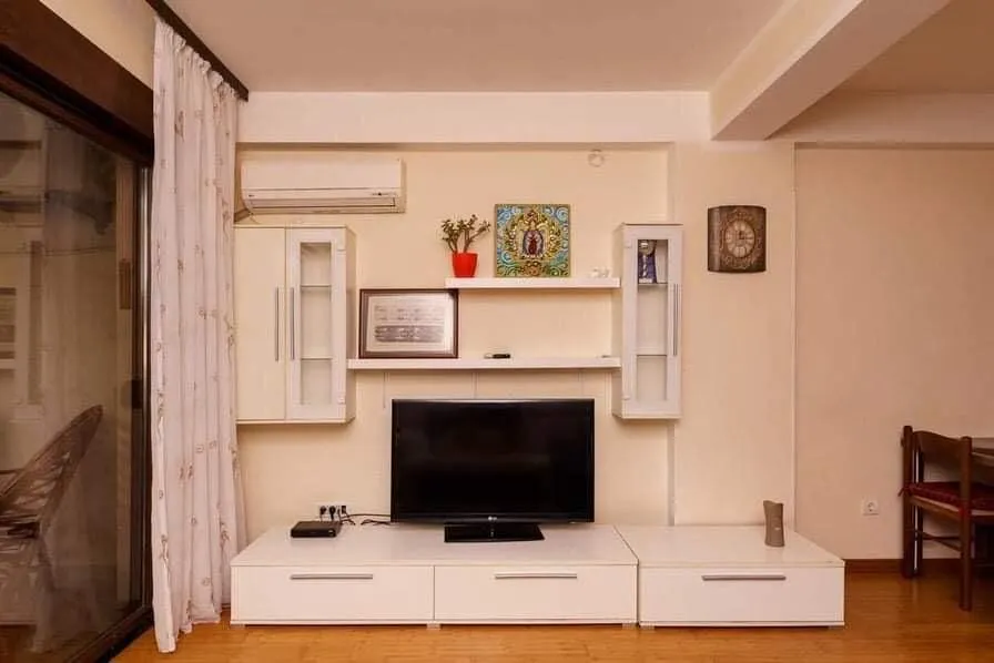 Dnevna soba u stanu u Budvi, Crna Gora: bela TV komoda, viseće police, klima-uređaj i izlaz na balkon.