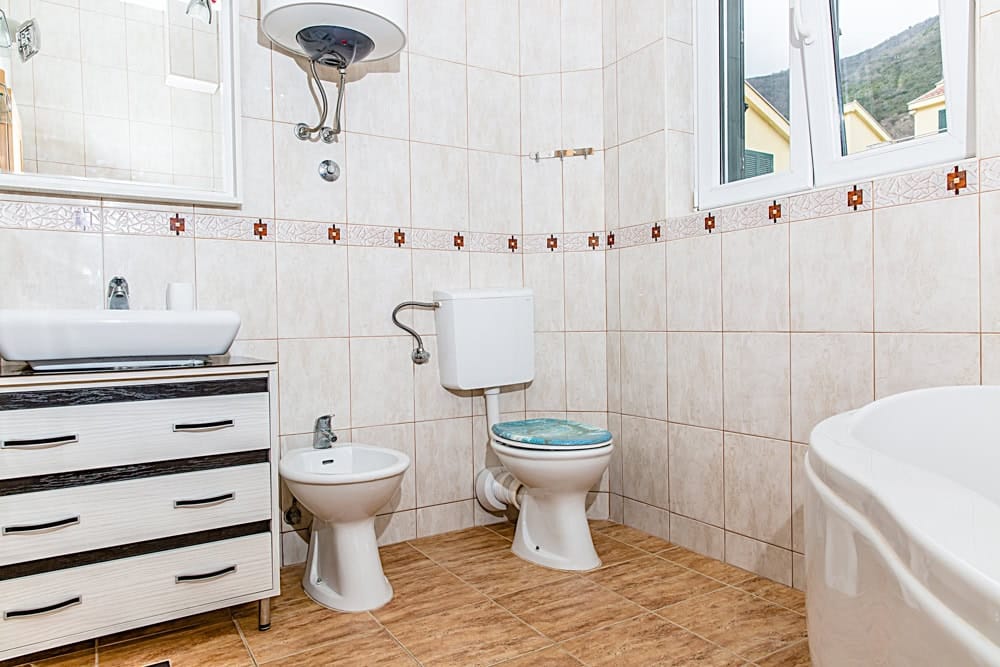 Jasna łazienka w mieszkaniu w Đenovici, Czarnogóra: szafka z umywalką, bidet, toaleta i widok na góry z okna.
