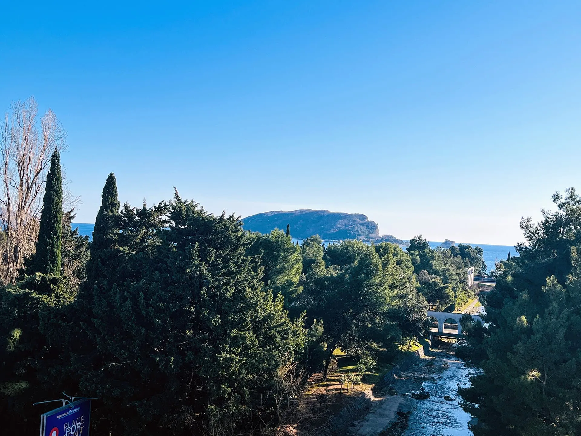 Panoramablick auf das Meer, die Berge und den Fluss in Budva, Montenegro, mit grünen Bäumen und einer Fußgängerbrücke im Vordergrund.