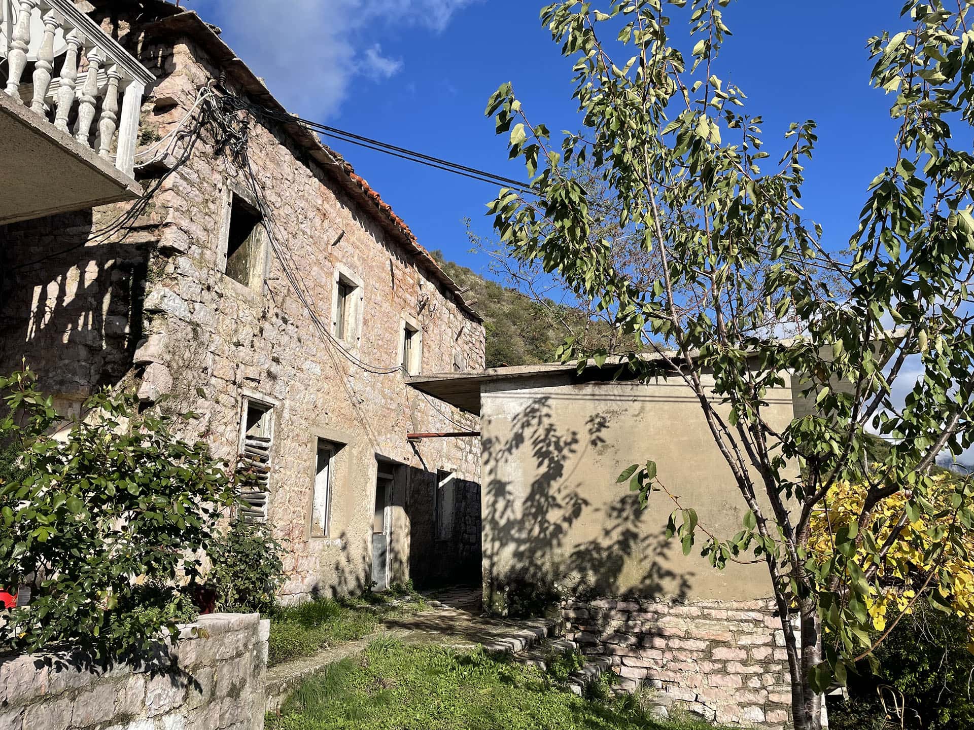 Ancienne maison en pierre à rénover sur un terrain à Kamenari, Monténégro, vue sur la façade et le terrain attenant.