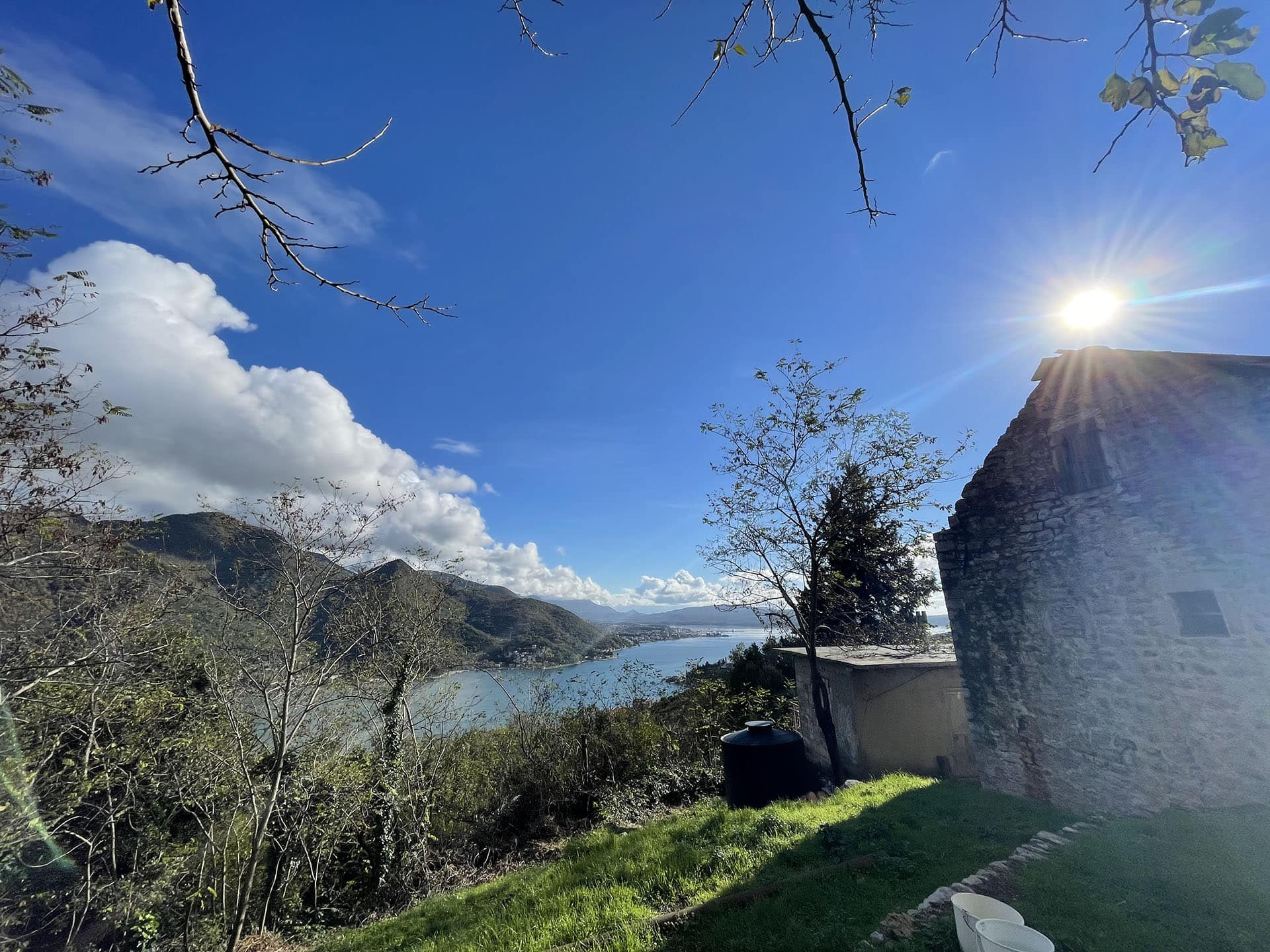 Vue panoramique sur la baie de Kotor et les montagnes depuis un terrain à Kamenari, Monténégro, avec une maison en pierre et un jardin.