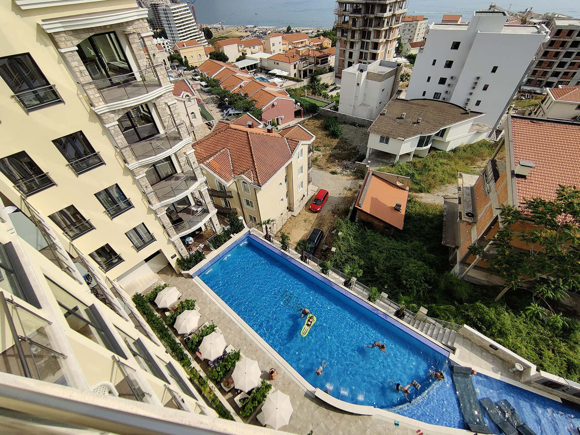 Top view of an outdoor pool at a residential complex in Bečići, Montenegro, with terraces and a cityscape.