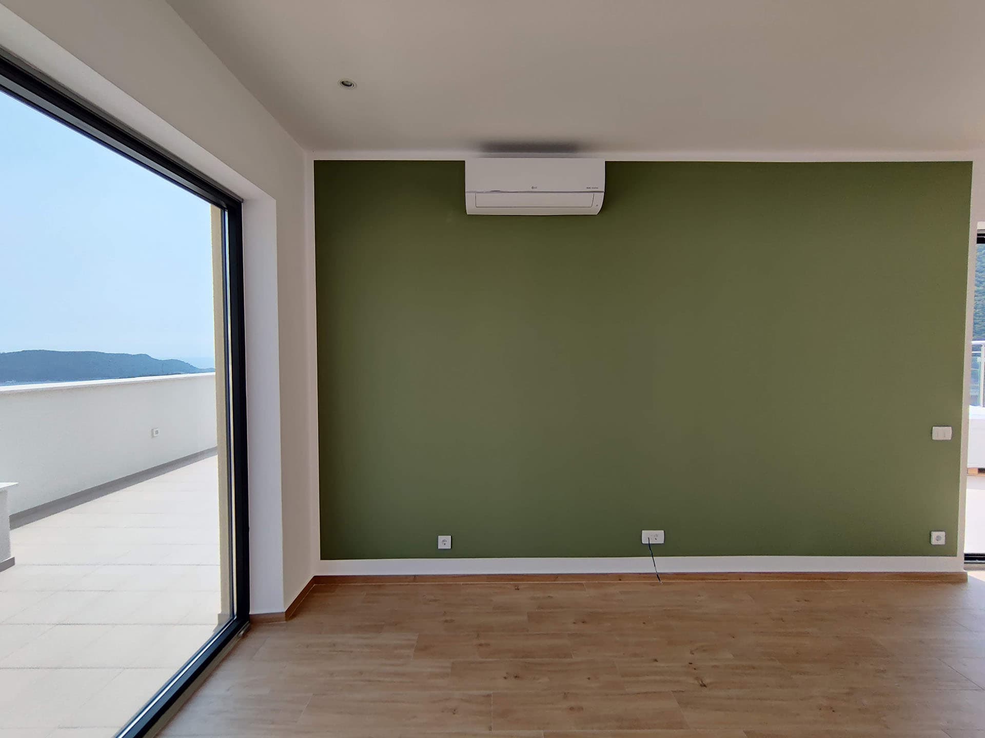 Living room with a panoramic window and sea view in an apartment, Becici, Montenegro: green wall, parquet flooring, and air conditioner.