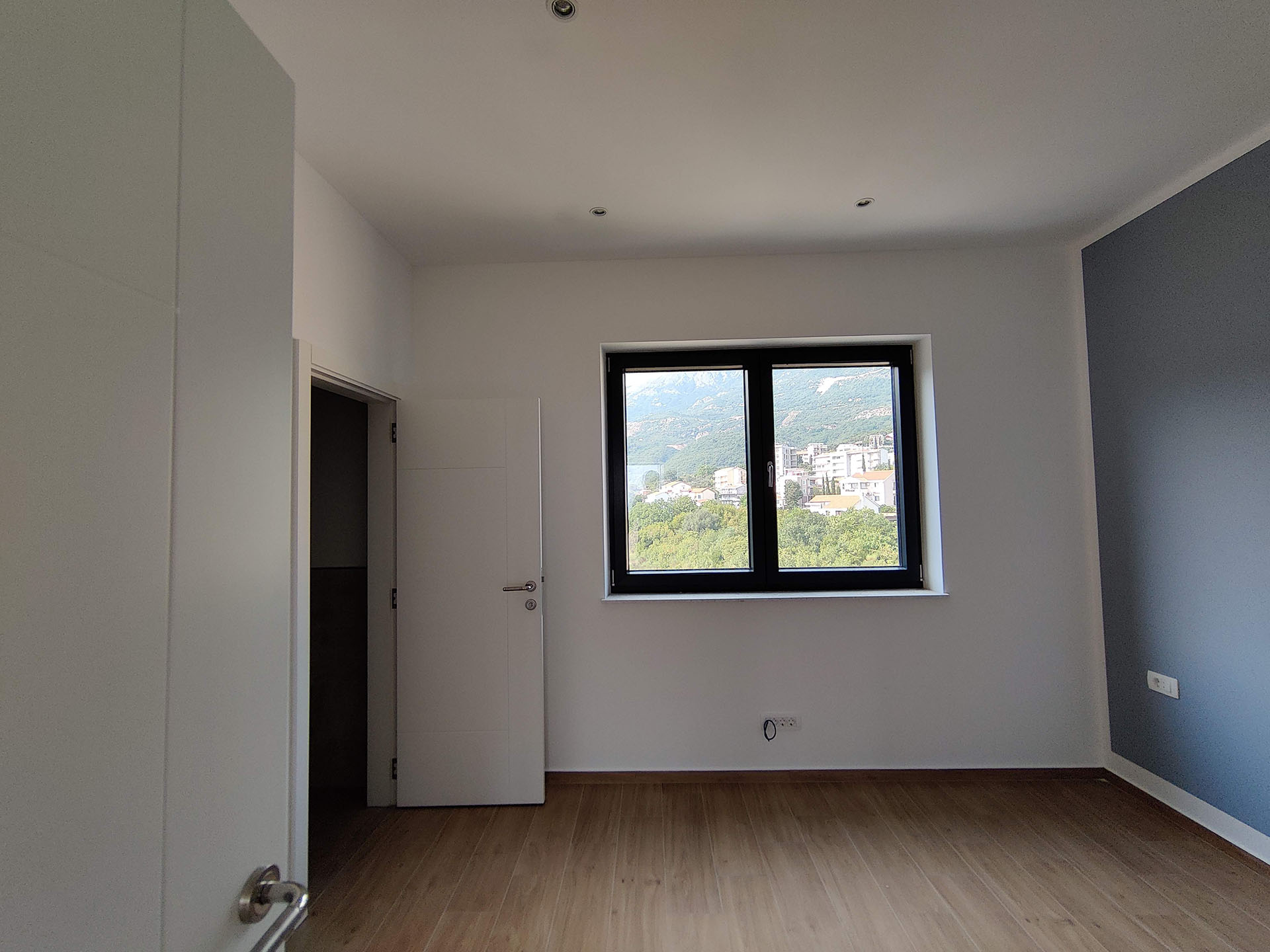 Bright bedroom in an apartment in Bečići, Montenegro: laminate flooring, white walls, an accent blue wall, and mountain views from the window.