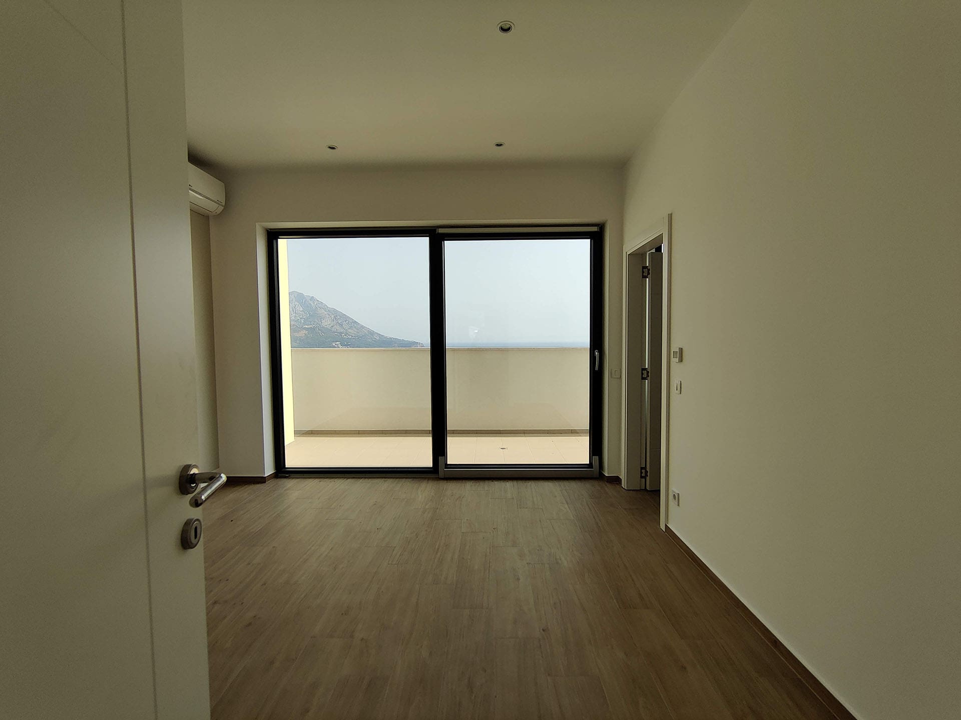 Bright living room in an apartment in Bečići, Montenegro, with a panoramic window, access to the terrace, and sea and mountain views.